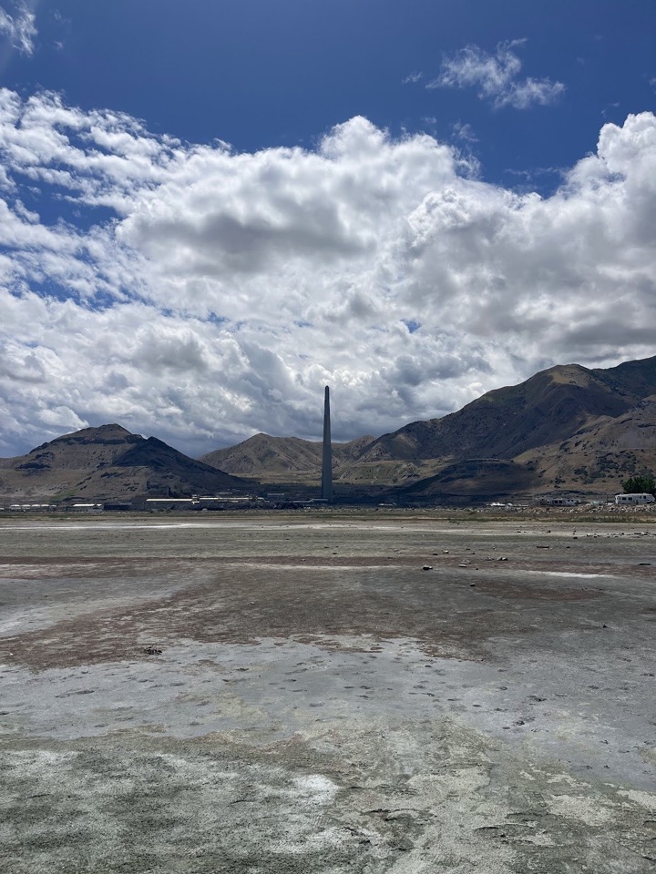 Salt flats under towering clouds, with mountains and a lone smokestack behind
